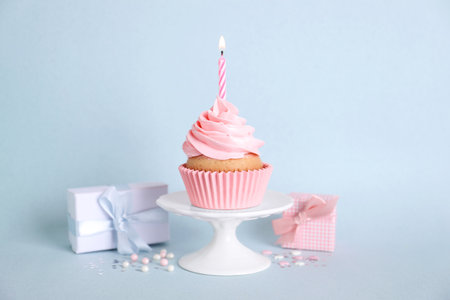 Delicious birthday cupcake with burning candle, sprinkles and gift boxes on light blue backgroundの写真素材