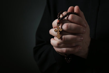 Priest with rosary beads praying on dark background, closeup. Space for textの写真素材