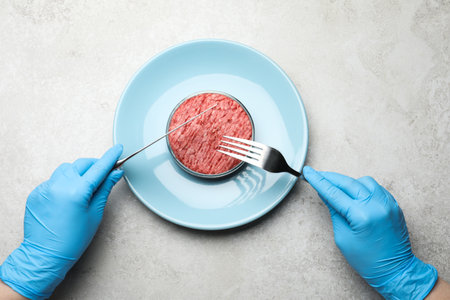 Scientist holding fork and knife over plate with minced cultured meat at light gray lab table, top viewの写真素材