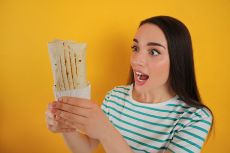 Emotional young woman holding tasty shawarma on yellow backgroundの写真素材