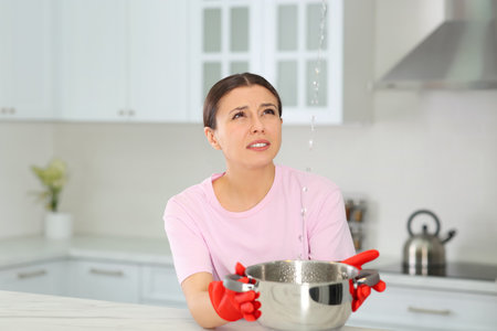 Young woman collecting leaking water from ceiling in kitchen. Time to call roof repair serviceの写真素材