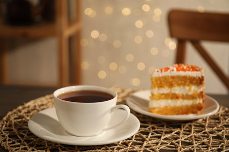 Hot tea in cup and piece of cake on wooden table against blurred lights indoorsの写真素材