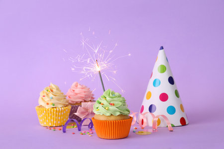 Birthday cupcakes with burning sparkler, party hat and gift box on violet backgroundの写真素材