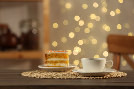 Hot drink in cup and piece of cake on wooden table against blurred lights indoorsの写真素材