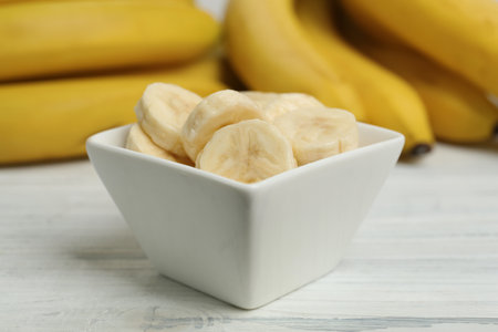 Bowl with cut bananas near whole fruits on white wooden table, closeupの写真素材