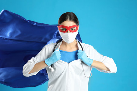 Doctor dressed as superhero posing on light blue background. Concept of medical workers fighting with pandemicの写真素材