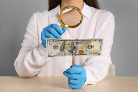 Expert authenticating 100 dollar banknote with magnifying glass at table on light gray background, closeup. fake money conceptの写真素材