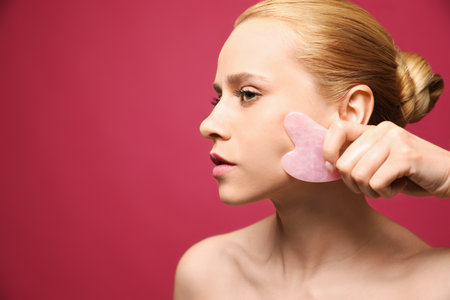 Beautiful young woman doing facial massage with gua sha tool on pink background, closeup. Space for textの写真素材