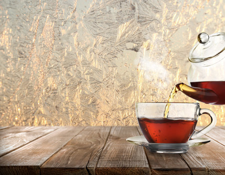 Pouring tea into glass cup on wooden table near window covered with frostの写真素材