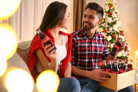 MYKOLAIV, UKRAINE - JANUARY 27, 2021: Young couple holding bottles of Coca-Cola in room decorated for Christmasのeditorial素材