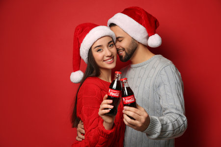 MYKOLAIV, UKRAINE - JANUARY 27, 2021: Young couple in Christmas hats holding bottles of Coca-Cola on red backgroundのeditorial素材
