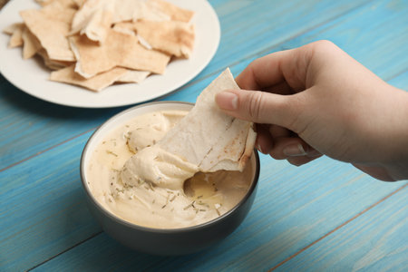 Woman dipping pita chip into hummus at turquoise wooden table, closeupの写真素材
