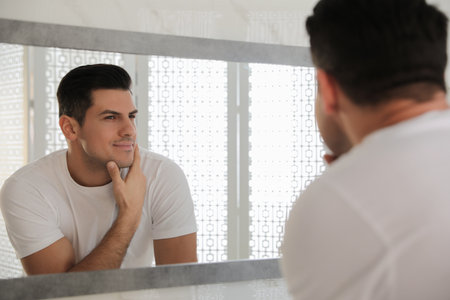 Handsome man touching his smooth face after shaving near mirror in bathroomの写真素材