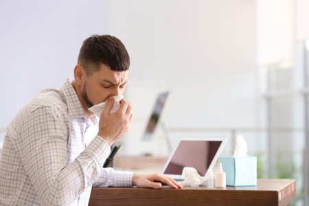 Ill man at table with nasal spray, drops and box of paper tissues in officeの写真素材