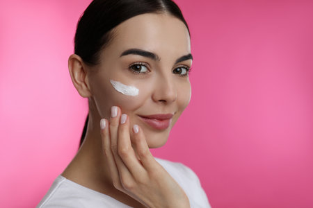 Young woman applying facial cream on pink backgroundの写真素材