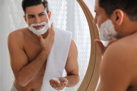 Handsome man applying shaving foam near mirror in bathroomの写真素材