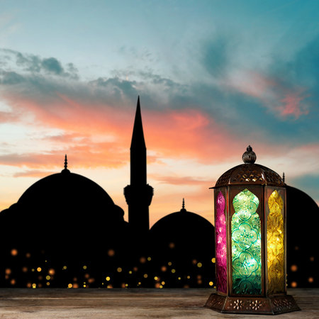 Decorative Arabic lantern on wooden surface and silhouette of mosque at sunset on background, space for textの写真素材