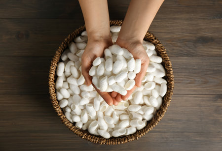 Woman holding white silk cocoons over bowl on wooden table, top viewの写真素材