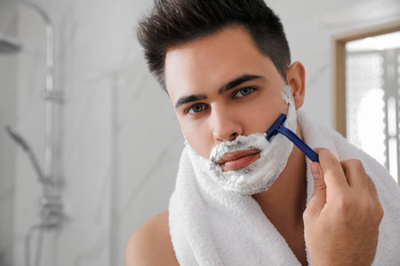 Handsome young man shaving with razor in bathroom, space for textの写真素材