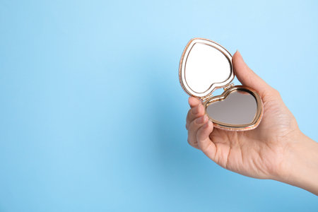 Woman holding stylish heart shaped cosmetic pocket mirror on light blue background, closeup. Space for textの写真素材