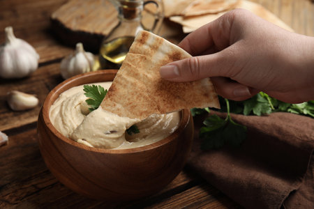 Woman dipping pita chip into hummus at wooden table, closeupの写真素材