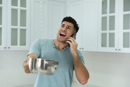 Emotional man calling roof repair service while collecting leaking water from ceiling in kitchenの写真素材