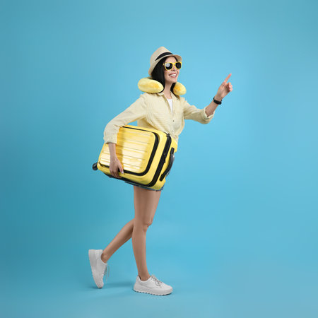 Happy female tourist with suitcase and travel pillow on light blue backgroundの写真素材
