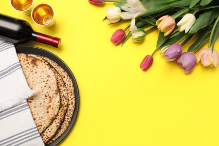 Tasty matzos, wine and fresh flowers on yellow background, space for text. Passover (Pesach)...