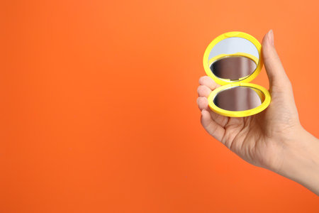 Woman holding stylish cosmetic pocket mirror on orange background, closeup. Space for textの写真素材