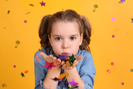 Adorable little girl blowing confetti on yellow background