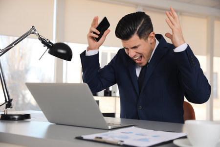 Emotional businessman with laptop and smartphone at table in officeの写真素材