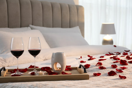 Tray with glasses of red wine, candles and rose petals on bed in roomの写真素材