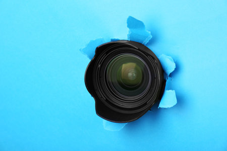Hidden camera lens through torn hole in light blue paperの写真素材