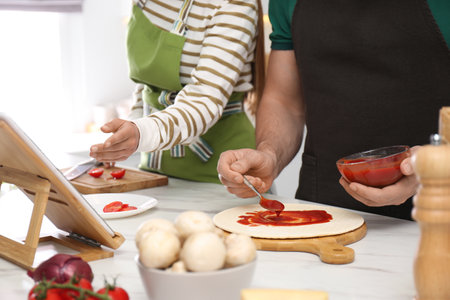 Couple making pizza together while watching online cooking course via tablet in kitchen, closeupの写真素材