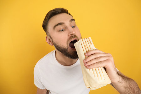 Young man eating delicious shawarma on yellow backgroundの写真素材