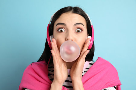 Fashionable young woman with headphones blowing bubblegum on light blue backgroundの写真素材