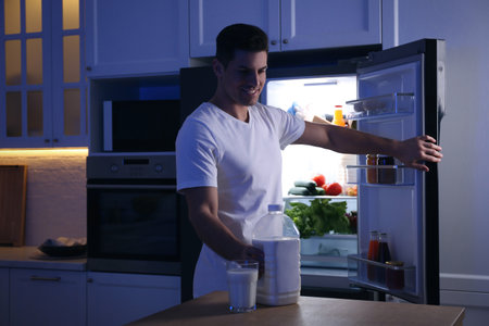 Man holding gallon bottle of milk and glass on wooden table in kitchen at nightの写真素材