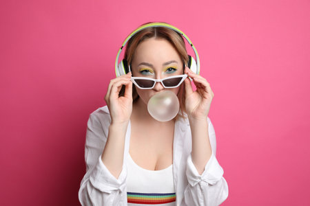 Fashionable young woman with bright makeup and headphones blowing bubblegum on pink backgroundの写真素材