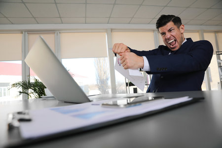 Emotional businessman with laptop at table in officeの写真素材