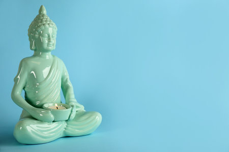 Beautiful ceramic Buddha sculpture with burning candle on light blue background. Space for textの写真素材
