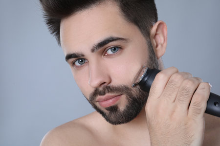 Handsome young man shaving with electric trimmer on gray background, closeupの写真素材