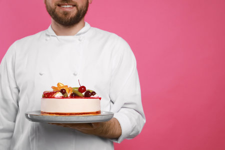 Happy professional confectioner in uniform holding delicious cake on pink background, closeup. Space for textの写真素材
