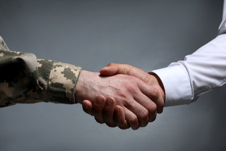 Soldier and businessman shaking hands against gray background, closeupの写真素材