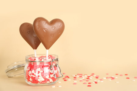 Chocolate heart shaped lollipops with sprinkles in glass jar on beige background. Space for textの写真素材