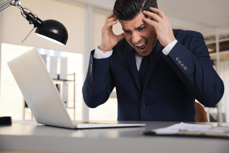 Emotional businessman with laptop at table in officeの写真素材