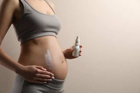 Pregnant woman applying cosmetic product on belly against beige background, closeup. Space for textの写真素材