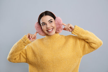 Beautiful young woman wearing earmuffs on light gray background