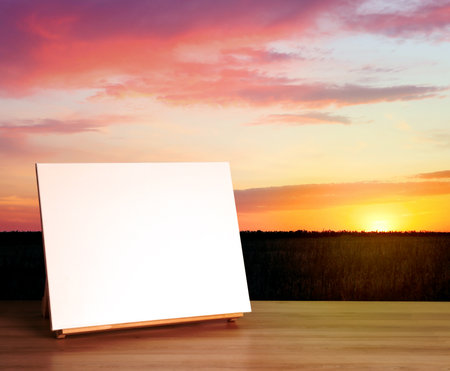 Wooden easel with blank canvas on table and beautiful field at sunset. Space for textの写真素材