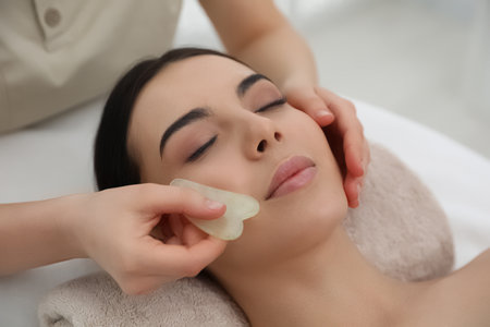 Young woman receiving facial massage with gua sha tool in beauty salonの写真素材