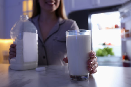 Young woman holding glass and gallon bottle of milk on white marble table in kitchen at night, closeupの写真素材